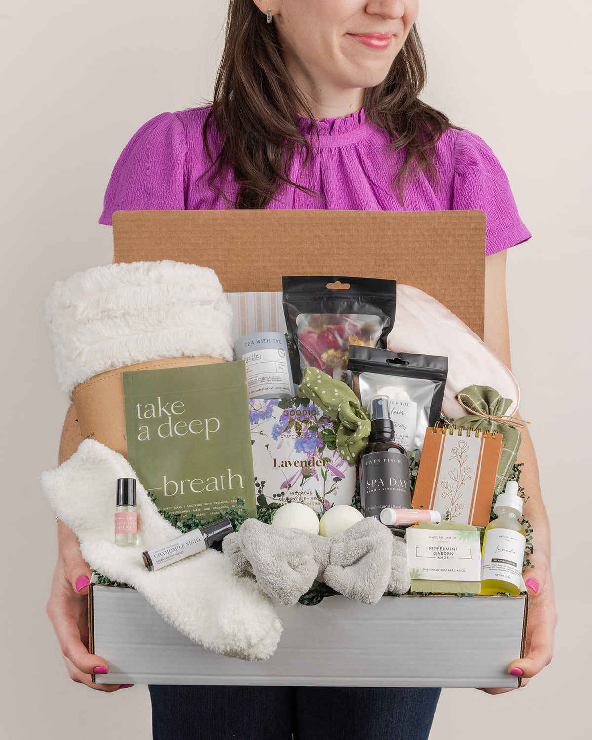 Person holding a gift box filled with a faux fur blanket, lavender chocolate, bath bombs, skincare, and spa accessories.