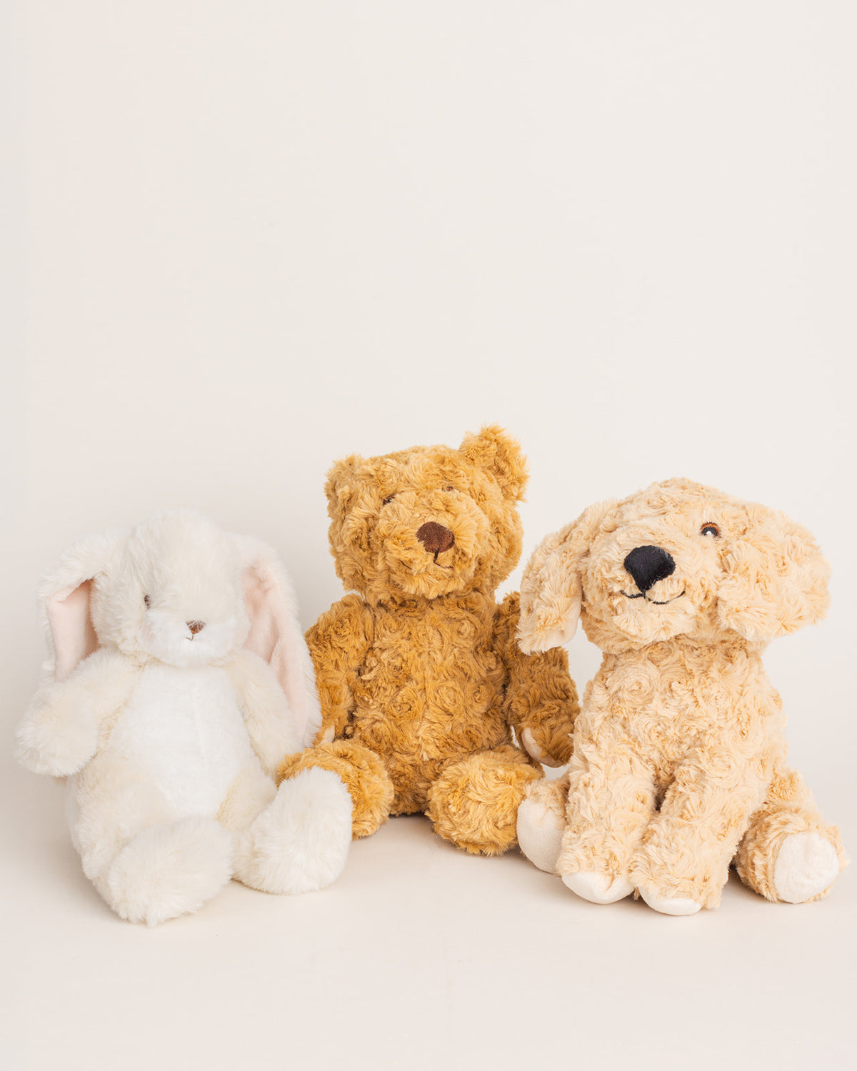 Three plush stuffed animals—a white bunny, a golden-brown teddy bear, and a tan puppy—sitting against a neutral background.