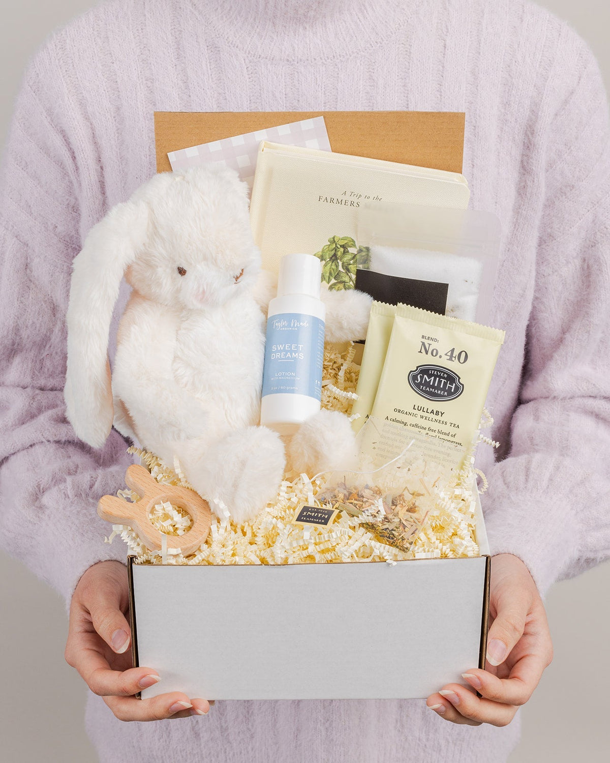 Deluxe baby gift box with a plush white bunny, wooden teether, organic tea, board book, and lotion in a cream-colored box.