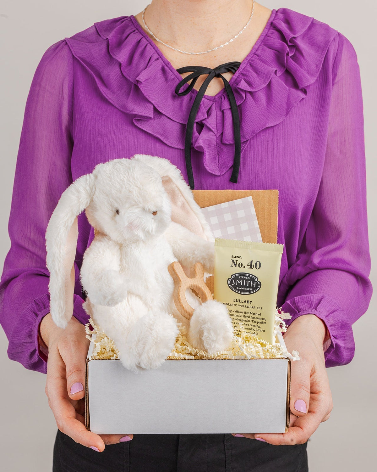 Welcome Baby gift box with a plush white bunny, wooden teething ring, and herbal tea, held by a person in a purple blouse.