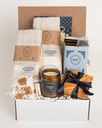 Welcome Home gift box with beige Turkish cotton towels, soy candle, matches, olive wood coasters, tea, and caramels.