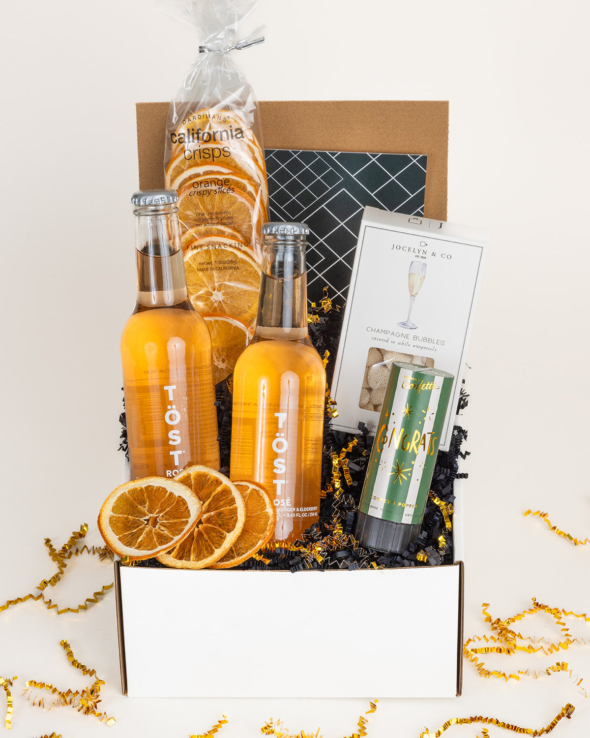 Gift box with two TÖST rosé bottles, orange crisps, champagne gummies, and a green confetti popper on black crinkle paper.