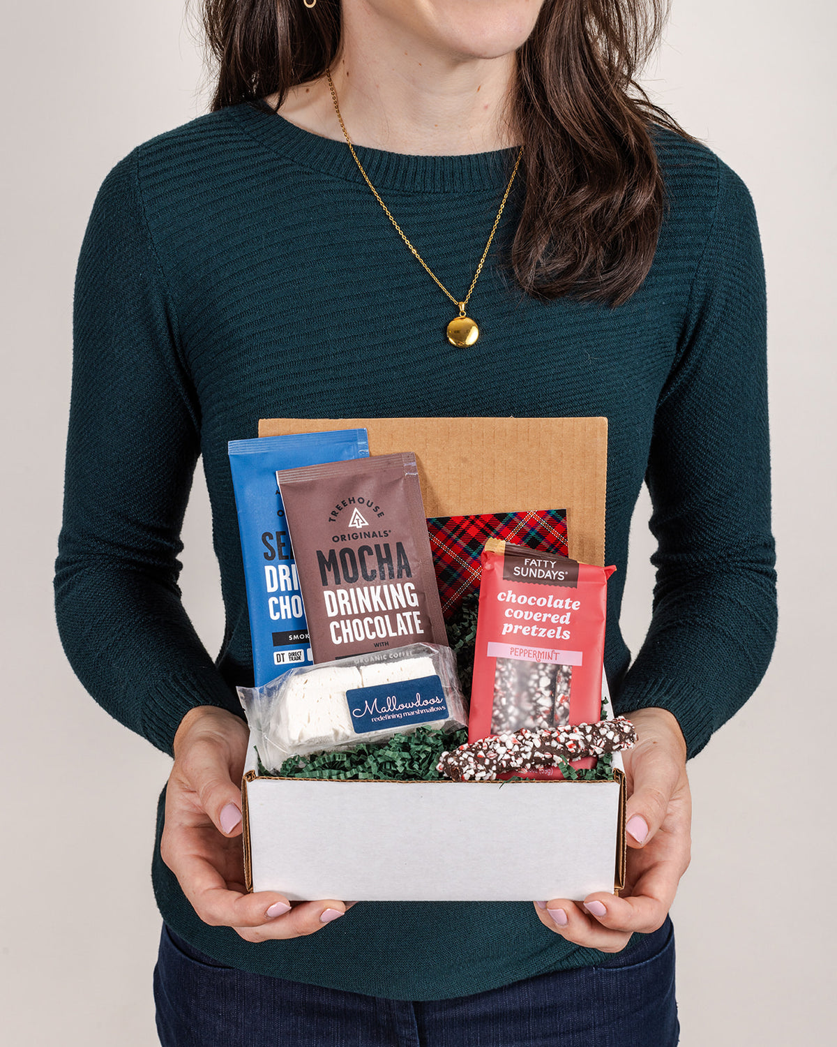 Person holding a gift box filled with drinking chocolate packets, handcrafted marshmallows, and chocolate-covered peppermint pretzels.