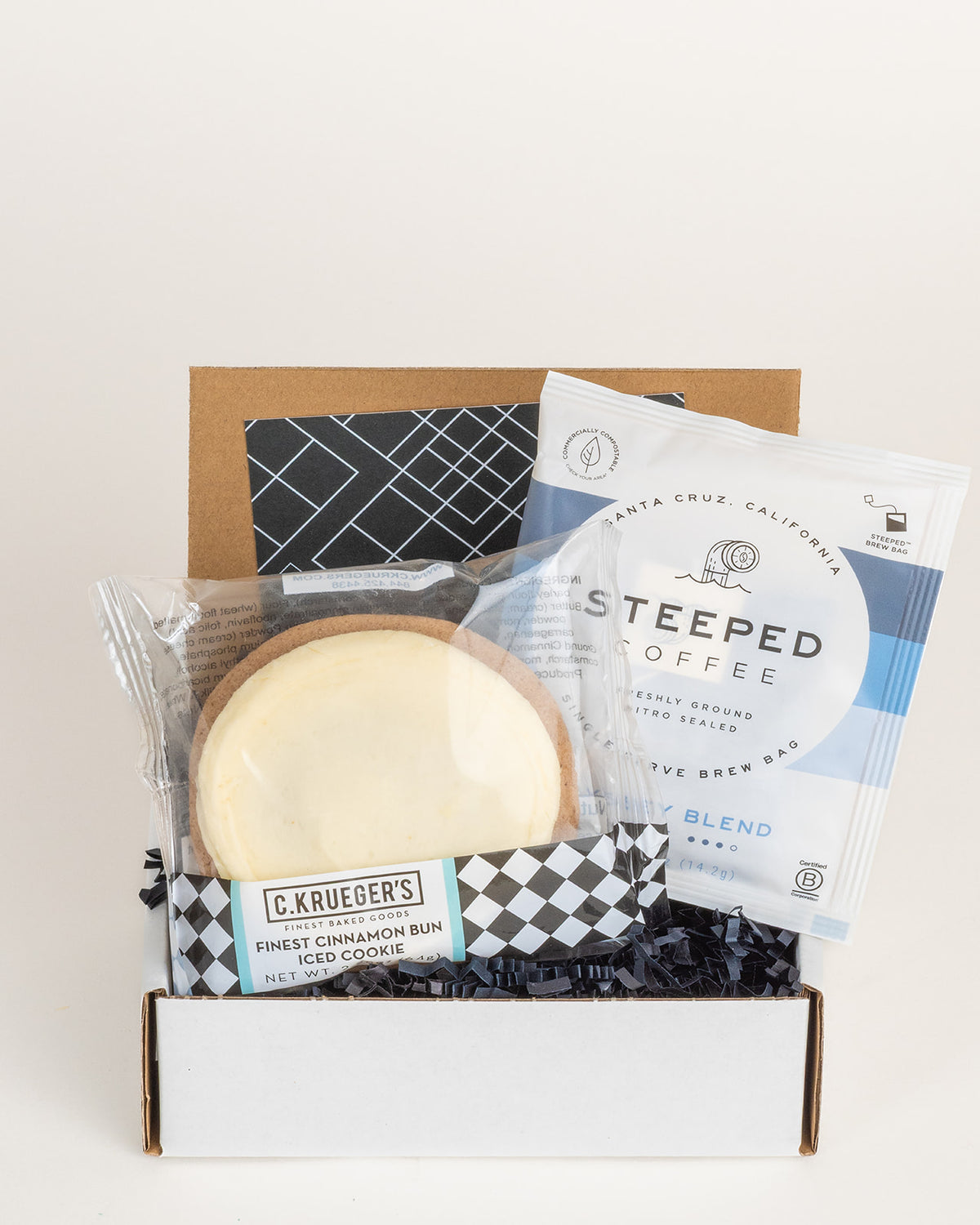Gift box containing a C. Krueger's iced cinnamon bun cookie and a Steeped Coffee brew bag nestled in black crinkle paper.