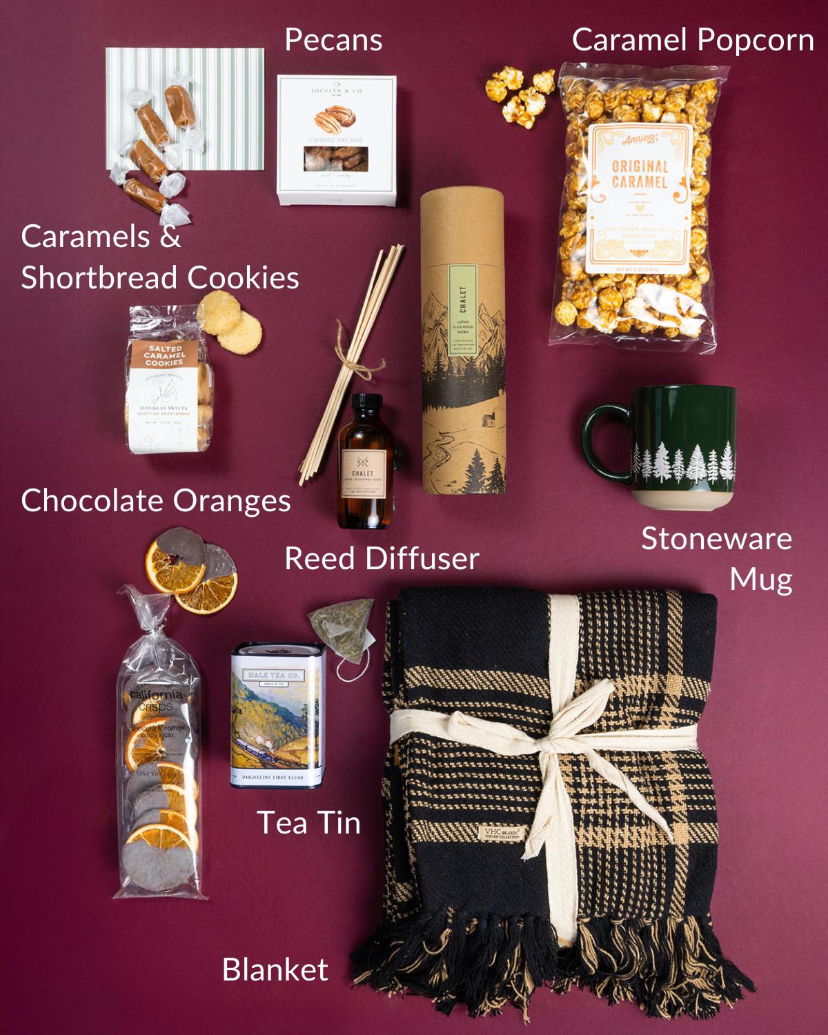 Cozy gift set with black plaid blanket, forest green stoneware mug, reed diffuser, gourmet snacks, and tea on burgundy.