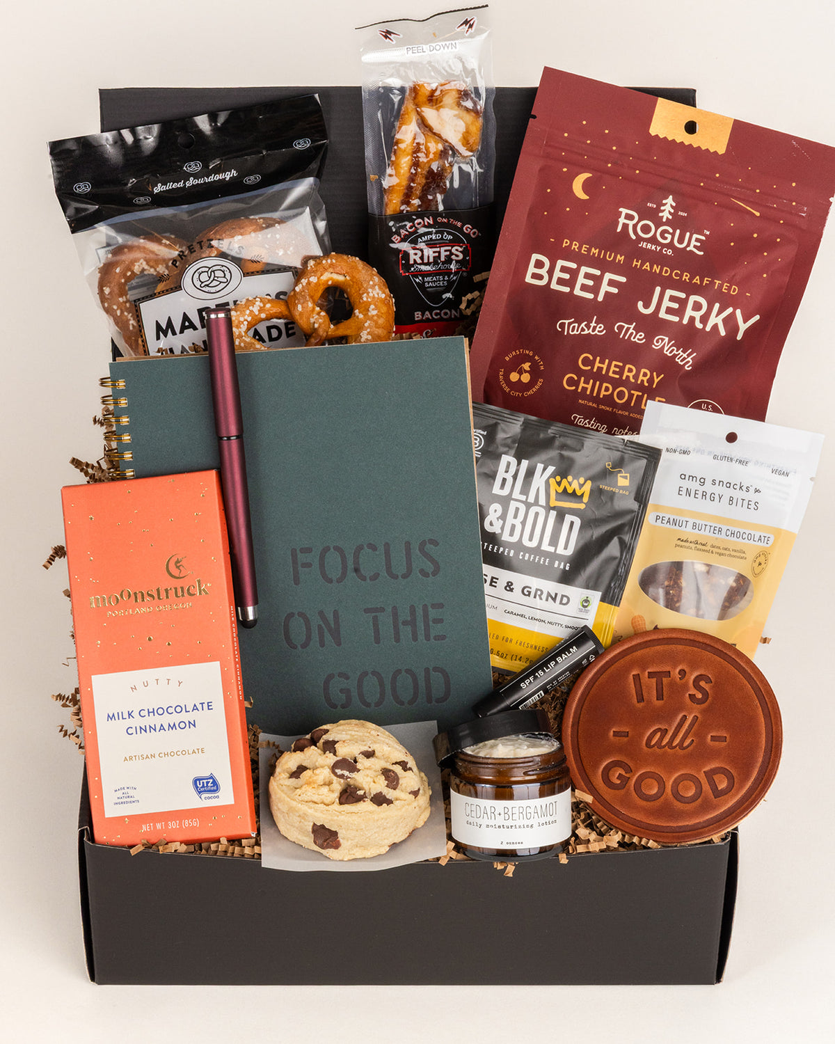 Focus On The Good gift box containing a green notebook, snacks, coffee, chocolate, and skincare items on brown paper filler.