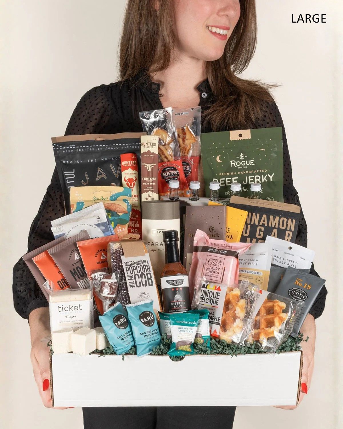 Woman holding a large white gift box filled with assorted premium snacks, jerky, chocolate, and gourmet pantry items.