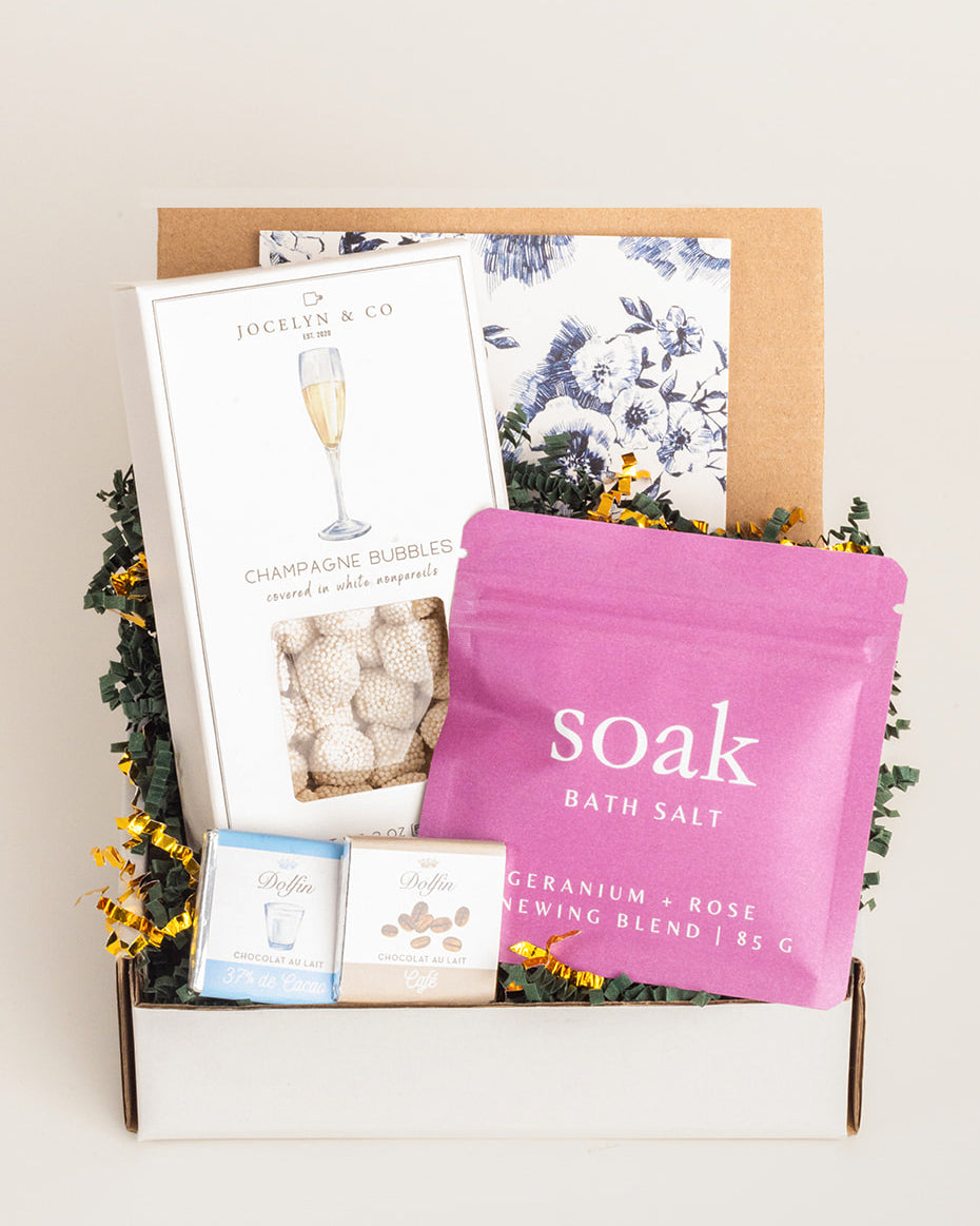 Gift box with champagne gummies, pink rose bath salts, two Dolfin chocolates, and a floral note on green paper shred.