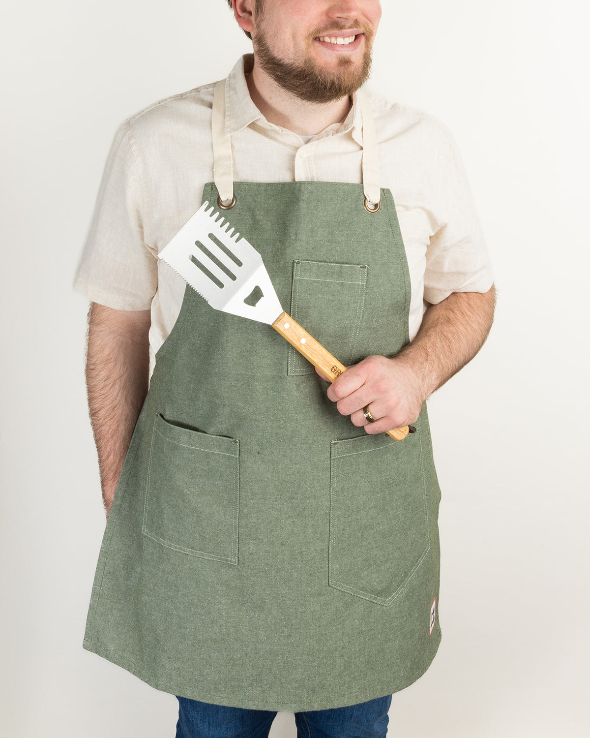 Man wearing a sage green canvas apron with pockets, holding a stainless steel grilling spatula with a wooden handle.