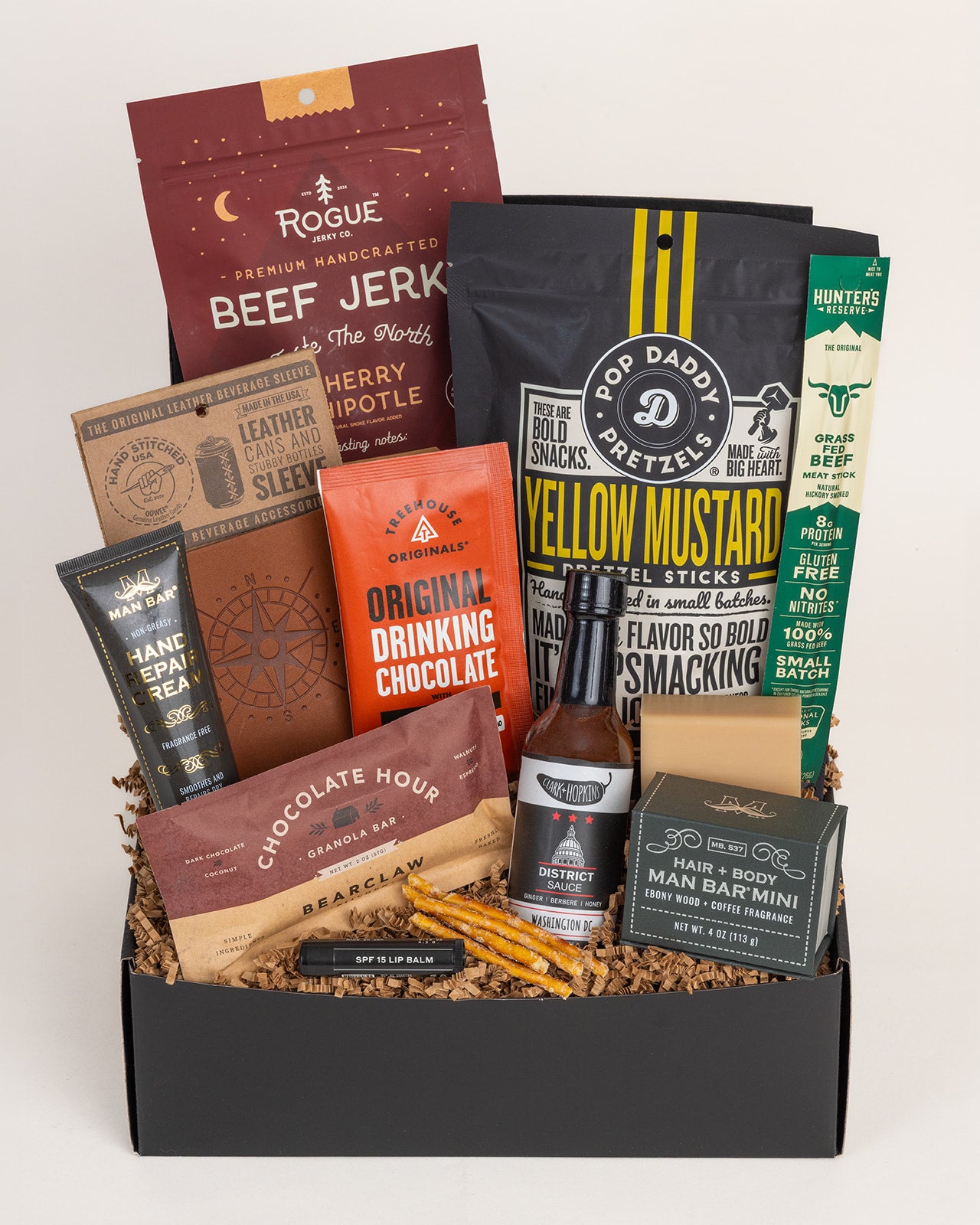 Rugged gift box featuring beef jerky, leather koozie, hot sauce, snacks, and grooming essentials in a black box with paper filler.