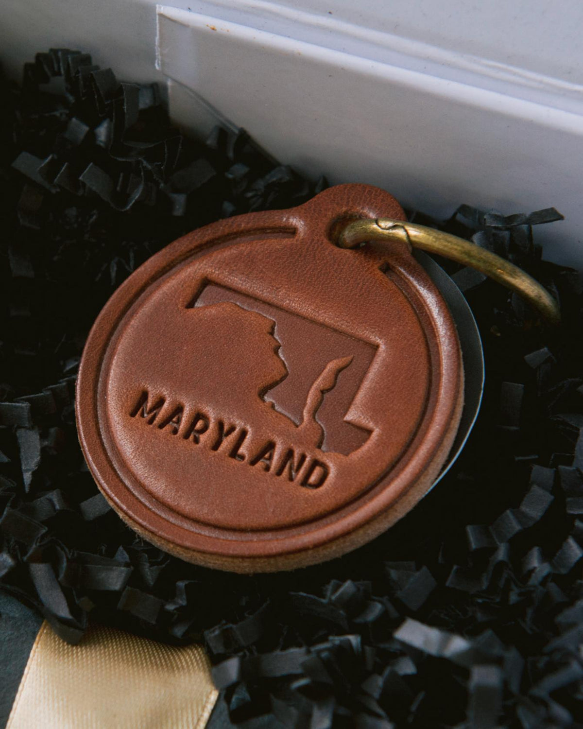 Round brown leather keychain embossed with a Maryland state map and text, nestled in black shredded paper gift box filler.