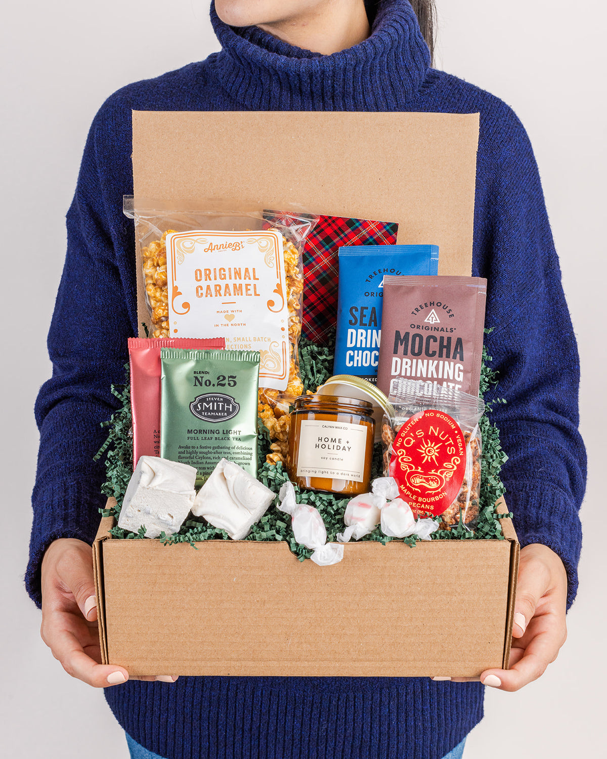 Person in a blue sweater holding a holiday gift box filled with caramel popcorn, drinking chocolate, tea, a candle, and treats.