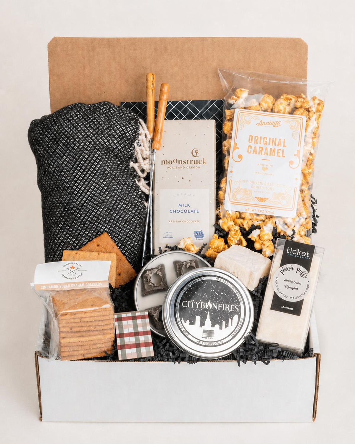 S'mores gift box featuring a black throw blanket, mini fire pit, roasting skewers, milk chocolate, marshmallows, and caramel popcorn.