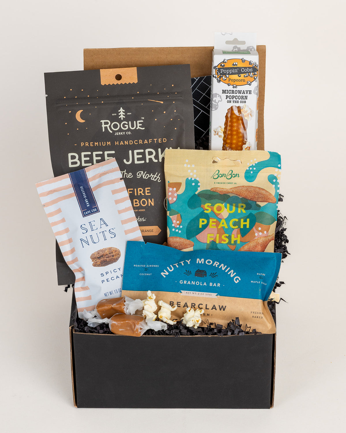 Gift box filled with gourmet snacks including beef jerky, popcorn, Swedish gummies, spicy pecans, and a granola bar on black crinkle paper.