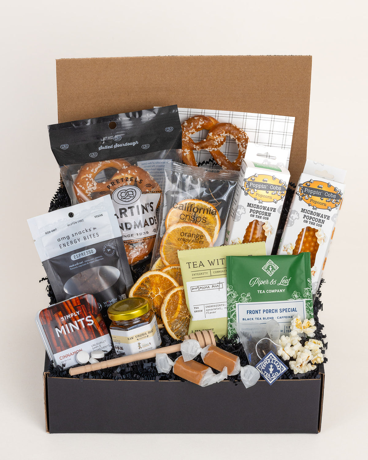 Gift box filled with assorted snacks including pretzels, popcorn on the cob, dried orange slices, tea, honey, and caramels.